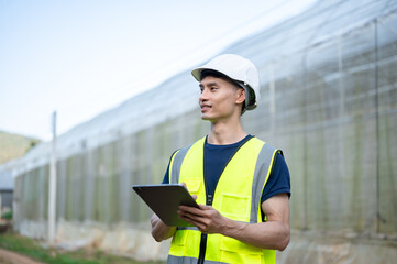 Asian engineer man wearing safety vest and hard hat helmet holding tablet aside greenhouse in farm.