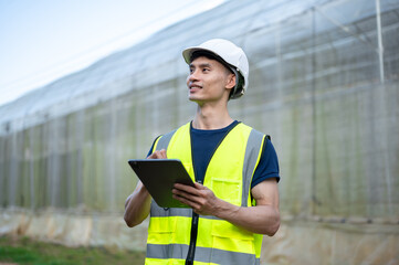 Asian engineer man wearings safety vest and hard hat helmet holding tablet standing aside greenhouse
