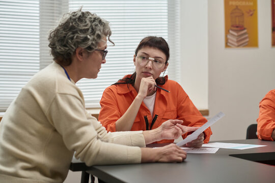 Middle aged Caucasian woman explaining document to young adult Caucasian woman in prison uniform during educational session in classroom setting, both seated at table, learning materials visible