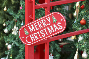 Red hanging sign saying Merry Christmas red metal frame near green pine tree decorated with gold and red ornament ball expressing joyful holiday celebration and festive season happiness atmosphere