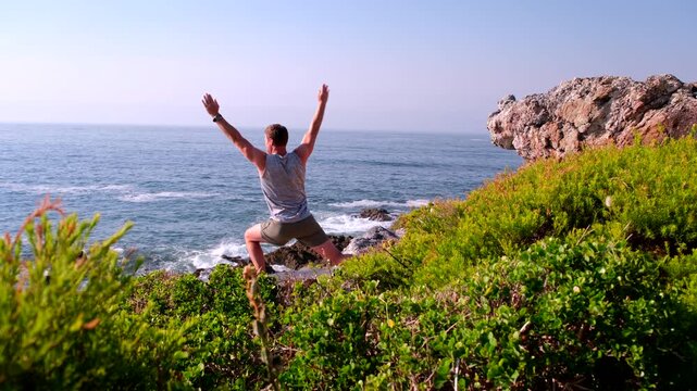 Yoga warrior poses by muscular man on sea cliff at sunrise overlooking ocean