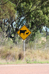 Yellow sign with black cow and sheep silhouettes warning drivers of livestock next to a country road with trees in the background