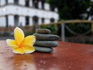 A peaceful Zen composition featuring a stack of four balanced smooth grey river stones next to a yellow plumeria flower on a red surface, with a soft-focus building in the background.