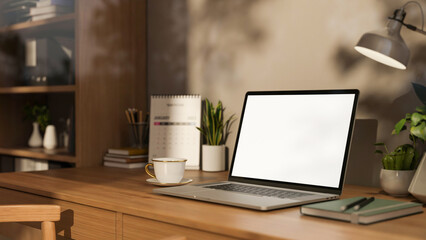 White screen laptop and coffee with lamp and calendar on wooden table aside bookshelf in study room.