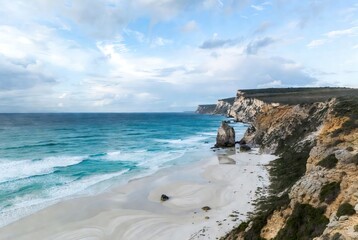 A serene coastal landscape featuring a white sandy beach, turquoise ocean, and rugged cliffs under a cloudy sky
