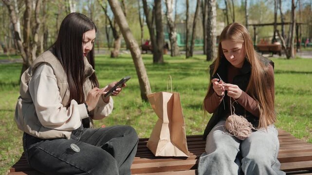 friends knitting and scrolling on bench, asian and white women share yarn and phone with paper bag between, needles and yarn in hands, relaxed park craft moment and light conversation