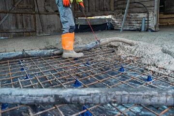 Concrete pouring for a building foundation is in progress, with a worker in protective gear managing the flow over rebar supported by chairs, ensuring structural integrity for the construction.