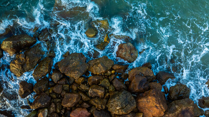 Angle drone shot capturing the beautiful contrast between the deep blue sea foam and the brown textured boulders along the shoreline, creating a natural abstract pattern.concept nature
