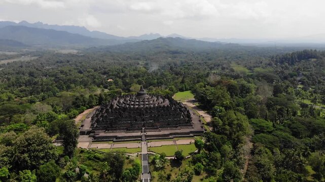Aerial Orbit Wide Shot of Borobudur Temple with Scenic Mountain Background Under Partly Cloudy Sky in 4K. Cinematic wide aerial drone view of the Borobudur Temple in Indonesia. The ancient Buddhist