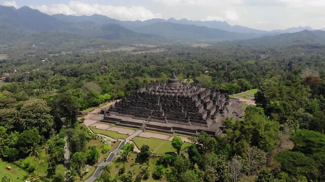 Aerial Orbit Wide Shot of Borobudur Temple with Scenic Mountain Background Under Partly Cloudy Sky in 4K. Cinematic wide aerial drone view of the Borobudur Temple in Indonesia. The ancient Buddhist