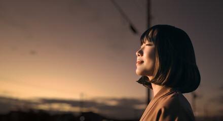 美しい夕焼け・朝焼け　夕日・朝日に照らされた空と日の出の光を浴びた背景　空を見上げ上を向きリラックスした表情の女性の横顔　日本・旅行・観光・アウトドア・リラクゼーションの背景　希望・安堵のイメージ