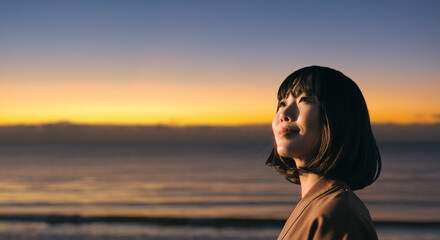 美しい夕焼け・朝焼け　夕日・朝日に照らされた海と日の出の光を浴びた背景　笑顔で空を見上げ上を向きリラックスした表情の女性の横顔　日本・旅行・観光・アウトドア・リラクゼーションの背景　希望・安堵のイメージ　ビジネスはじまり・出勤・転職・就職