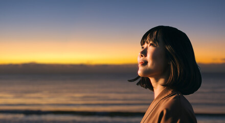 美しい夕焼け・朝焼け　夕日・朝日に照らされた海と日の出の光を浴びた背景　笑顔で空を見上げ上を向きリラックスした表情の女性の横顔　日本・旅行・観光・アウトドア・リラクゼーションの背景　希望・安堵のイメージ　ビジネスはじまり・出勤・転職・就職