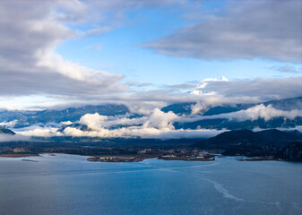 Fototapeta premium Snowy Mountain Peak Rises Over Cloudy Fjord With Calm Water in Squamish Area