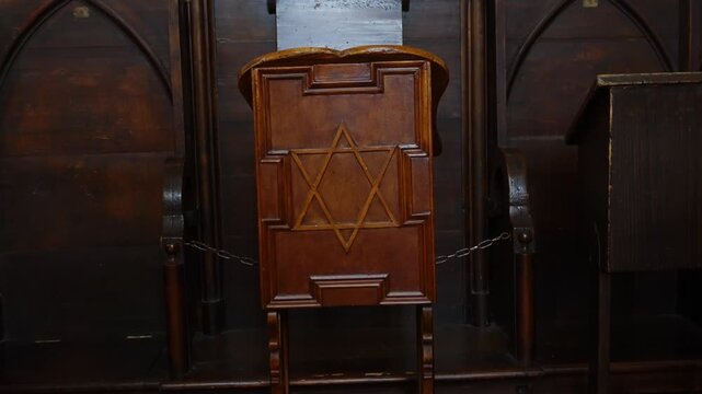 Dimly lit synagogue interior showing carved wooden chair and dark architecture with solemn atmosphere