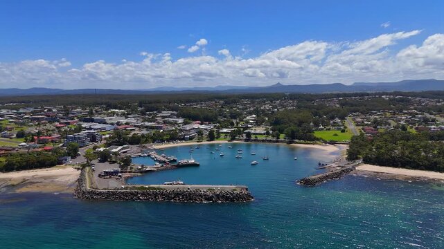 Drone angled approach to Ulladulla harbour mouth opening to the Pacific Ocean, blue sky with soft clouds over homes and buildings