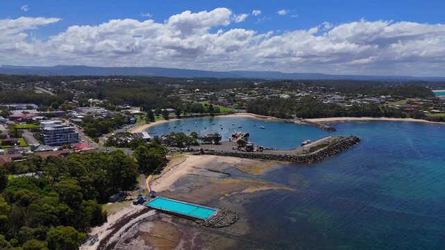 Aerial tracking right orbit of Ulladulla waterfront with marina, boats, and town buildings, open bath pools on edge