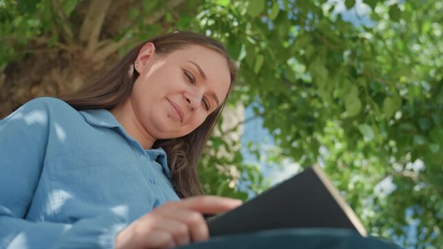 woman reviews manuscript, woman examines draft under trees, woman thoughtfully inspects written work beneath leaves, gentle woman studies her document amidst shaded outdoor setting with warmth