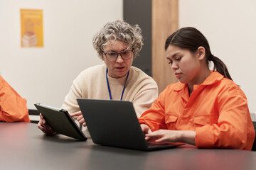 Middle aged Caucasian woman mentoring young adult woman using laptop in classroom setting, both wearing orange uniforms, participating in prison education program