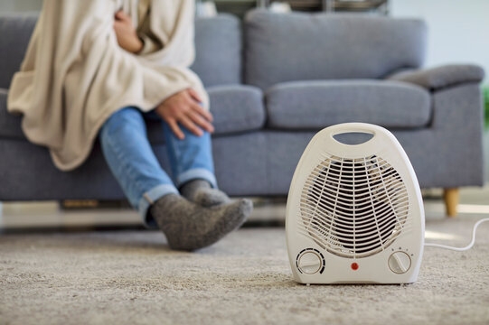 Man with blanket freezing near heater at home. Sitting on sofa in chilly cold room, he warming up on a portable electric heater to add warmth and comfort. Concept winter heating problem.