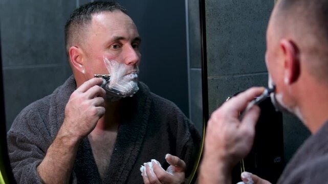 Hispanic bearded handsome man shaving using disposable razor in bathroom looking at mirror. Daily morning routine male hygienic beauty procedure. Guy shaves beard and moustache
