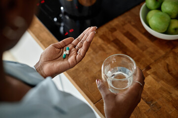 Black woman holding glass of water in one hand and several capsules in other hand, standing in kitchen near bowl of green apples, preparing to take medication