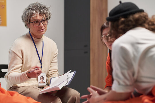 Middle aged Caucasian woman with curly gray hair holding open notebook, engaging in discussion with two young adult women in orange uniforms during prison therapy session