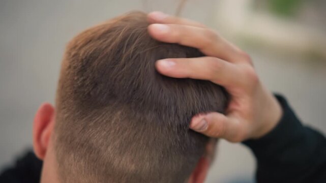 young man checking hair crown and scalp from top view, fingers smoothing short brown hair, closeup of roots and texture, mirror and styling preparation implied, quiet pre-shoot routine, natural light