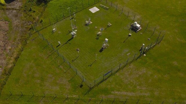 Aerial summer view of a fenced weather monitoring station with scientific instruments on a green field in Estonia captured on a sunny day.