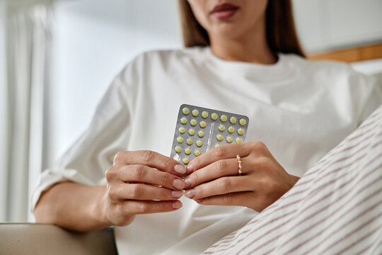 Young adult Caucasian woman holding blister pack of oral contraceptive pills with both hands, sitting indoors, face partially visible, focusing on medication packaging