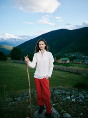 Fototapeta premium Nature, countryside, girl, hiking, outdoors, mountains, landscape, walking stick, rural, scenery describe a girl standing on rocky ground in a white knit shirt and red pants under a blue sky in wide