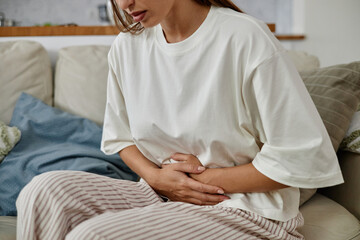 Young adult Caucasian woman sitting on sofa holding stomach with both hands appearing to experience...