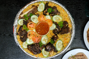 A top-down view of a Mandi platter featuring roasted lamb, yellow basmati rice, sliced cucumbers, and tomatoes on a foil tray with a central red sauce.