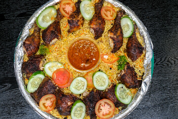 Top view of a Chicken Mandi platter featuring roasted chicken pieces, yellow basmati rice, cucumber, and tomato slices. A red sauce cup is centered on the foil-lined round tray.