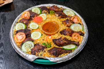 Top view of a Chicken Mandi platter featuring roasted chicken pieces, yellow basmati rice, cucumber, and tomato slices. A red sauce cup is centered on the foil-lined round tray.