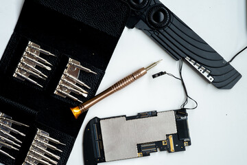 A disassembled smartphone internal motherboard and ribbon cables placed on a white surface next to a gold-toned precision screwdriver and an open black leatherette bit set.