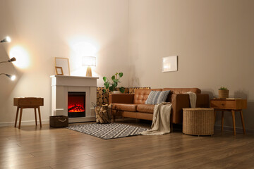 Interior of living room with sofa, glowing lamps, bedside tables and firewood near fireplace in...
