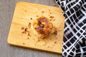 Traditional Indonesian bakpia pastries on a rustic wooden board with scattered crumbs. High-angle food photography featuring two golden-brown flaky treats and a checkered cloth background.