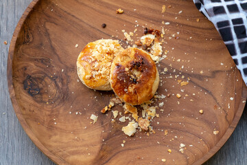 Traditional Indonesian bakpia pastry on a round wooden plate with scattered crumbs. High-angle food photography of a single flaky, golden treat with a checkered cloth detail.