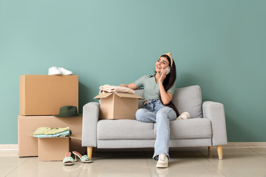 Young woman with wardrobe boxes talking by mobile phone on sofa near green wall