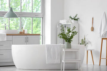 Interior of stylish bathroom with bathtub, shelving unit, commode and green blossoming branches in vases near window