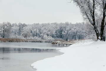 Calm winter landscape showing a frozen lake, snowy riverbank, and frost-covered trees creating a quiet, peaceful natural scene.