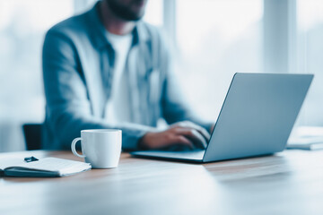 Man working on laptop at desk with coffee cup and notebook, focused on task in bright modern office environment