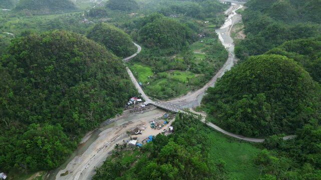 Peaceful aerial flyover of riverside barangay town, bridge, and winding roads amid lush tropical valley at Hicming, Catanduanes, Philippines