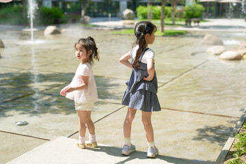 Two little girls standing in front of a fountain. One is wearing a pink shirt and the other is wearing a blue dress