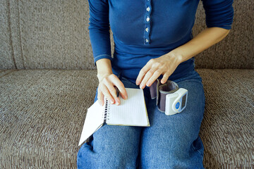 Woman recording blood pressure readings in a notebook with a digital monitor beside her. Symbolizes health data journaling, medical appointment preparation, and symptom tracking discipline.