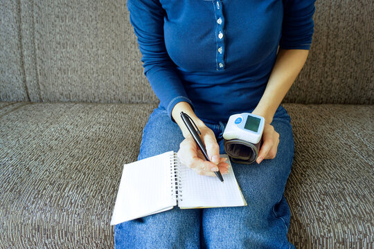 Young woman holding a blood pressure monitor and writing health observations in a notebook. Symbolizes health record keeping, cardiovascular risk awareness, and proactive disease prevention.
