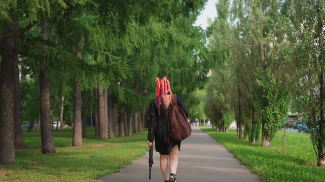 redhaired artist heading to performance space, caucasian performer carrying gear on scenic outdoor path, lightskinned musician with red dreadlocks strolls towards musical engagement outdoors