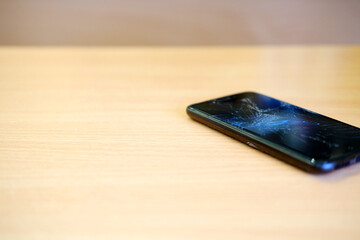 Broken smartphone lies on a wooden school desk, showcasing a cracked screen and reflecting light, symbolizing the challenges of student life and technology reliance