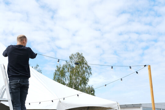 Worker prepares a festive tent for celebration, hanging decorative lanterns against a backdrop of blue sky and fluffy clouds, creating a joyful atmosphere for an outdoor event